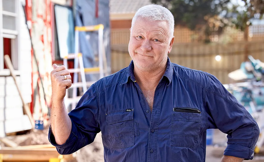 Man in a blue shirt standing outdoors, smiling, in front of a construction site.