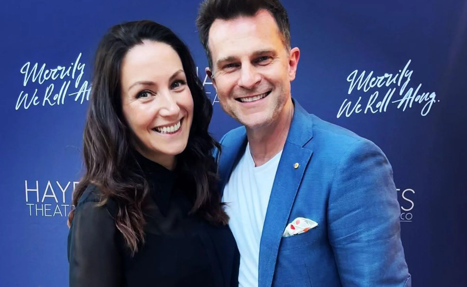 Couple smiling at the "Merrily We Roll Along" event backdrop.