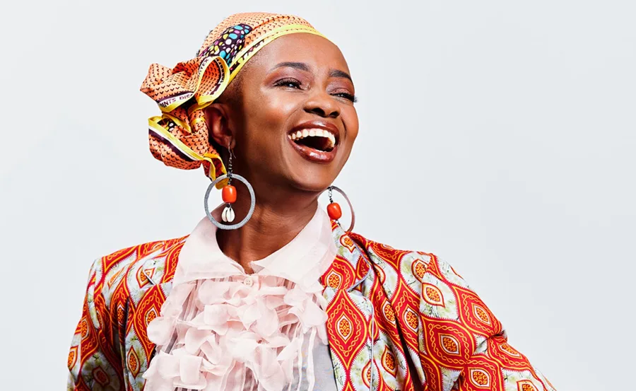 A woman wearing vibrant patterned clothes and a headscarf smiles broadly with large hoop earrings.