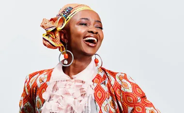 A woman wearing vibrant patterned clothes and a headscarf smiles broadly with large hoop earrings.