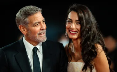 Couple smiling on red carpet, man in suit with gray hair, woman with long dark hair in sparkling dress.