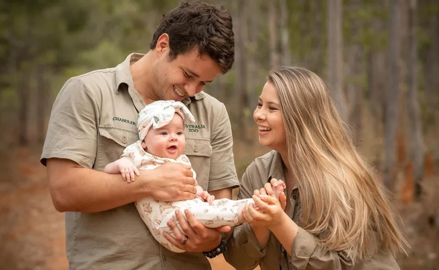 Couple joyfully holding a smiling baby outdoors, wearing matching khaki outfits with "Australia Zoo" logos.