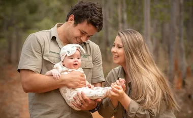 Couple joyfully holding a smiling baby outdoors, wearing matching khaki outfits with "Australia Zoo" logos.