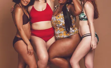 Four women of diverse body types wearing colorful swimsuits, embracing and smiling against a neutral background.
