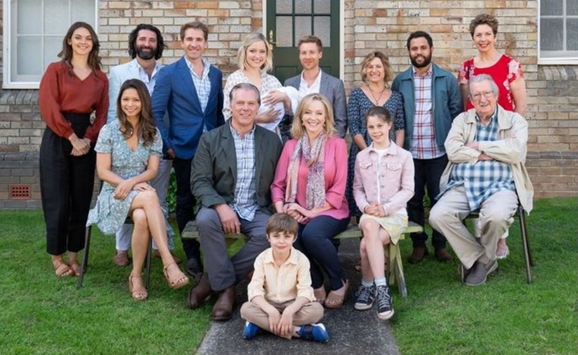 A group photo of "Back to the Rafters" cast standing and seated outside a brick house, smiling at the camera.
