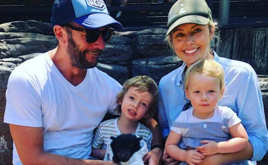 Family sitting together outdoors, smiling. Father and son hold a small animal, mother holds daughter.