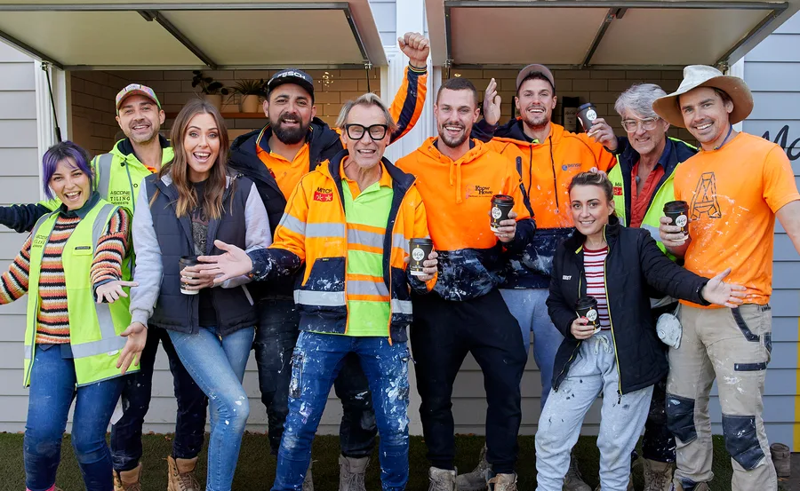 Group of smiling construction workers and team members in casual clothing, holding coffee cups, in front of a house.