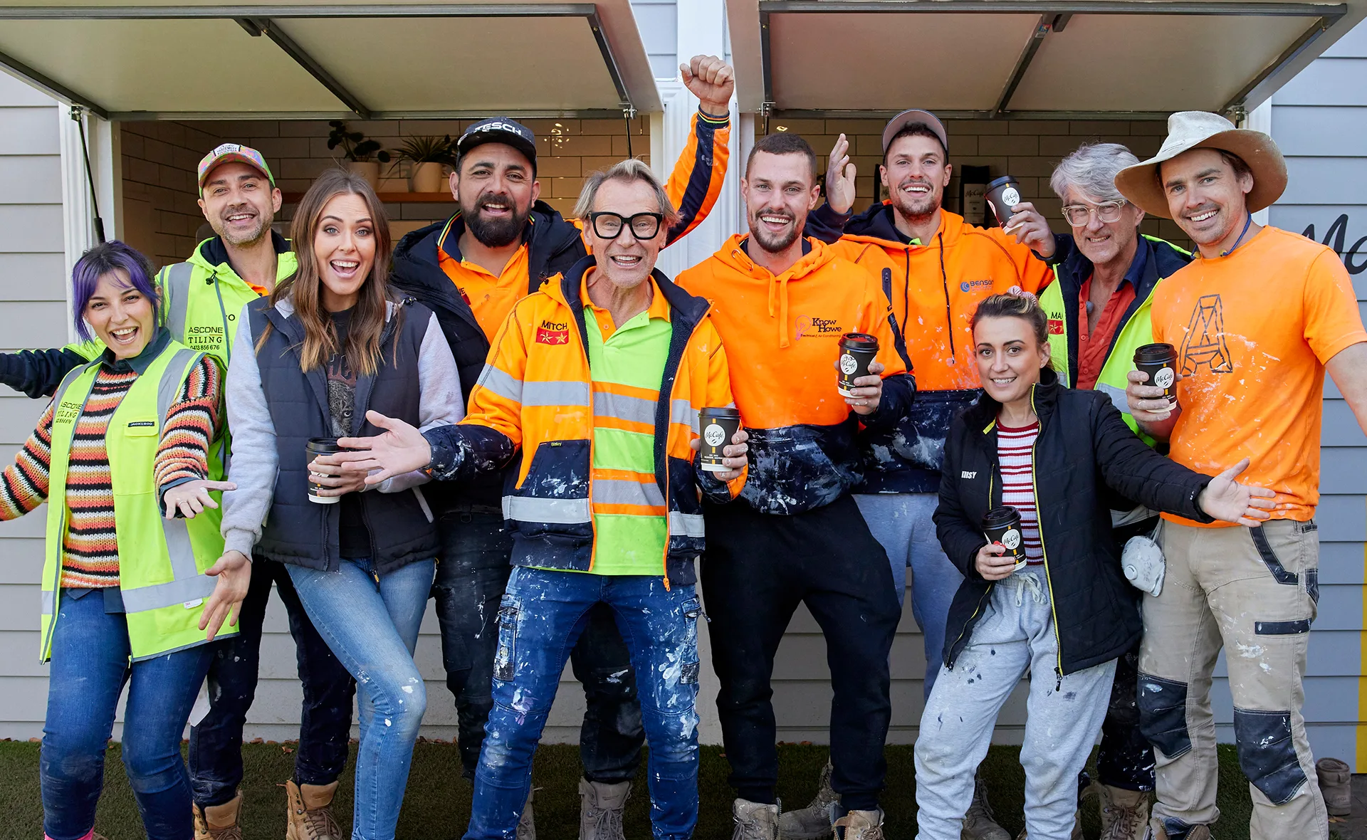 Group of smiling construction workers and team members in casual clothing, holding coffee cups, in front of a house.
