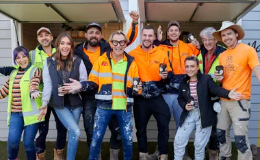 Group of smiling construction workers and team members in casual clothing, holding coffee cups, in front of a house.