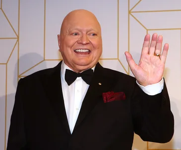 A smiling man in a tuxedo waves at an event with a patterned geometric background.