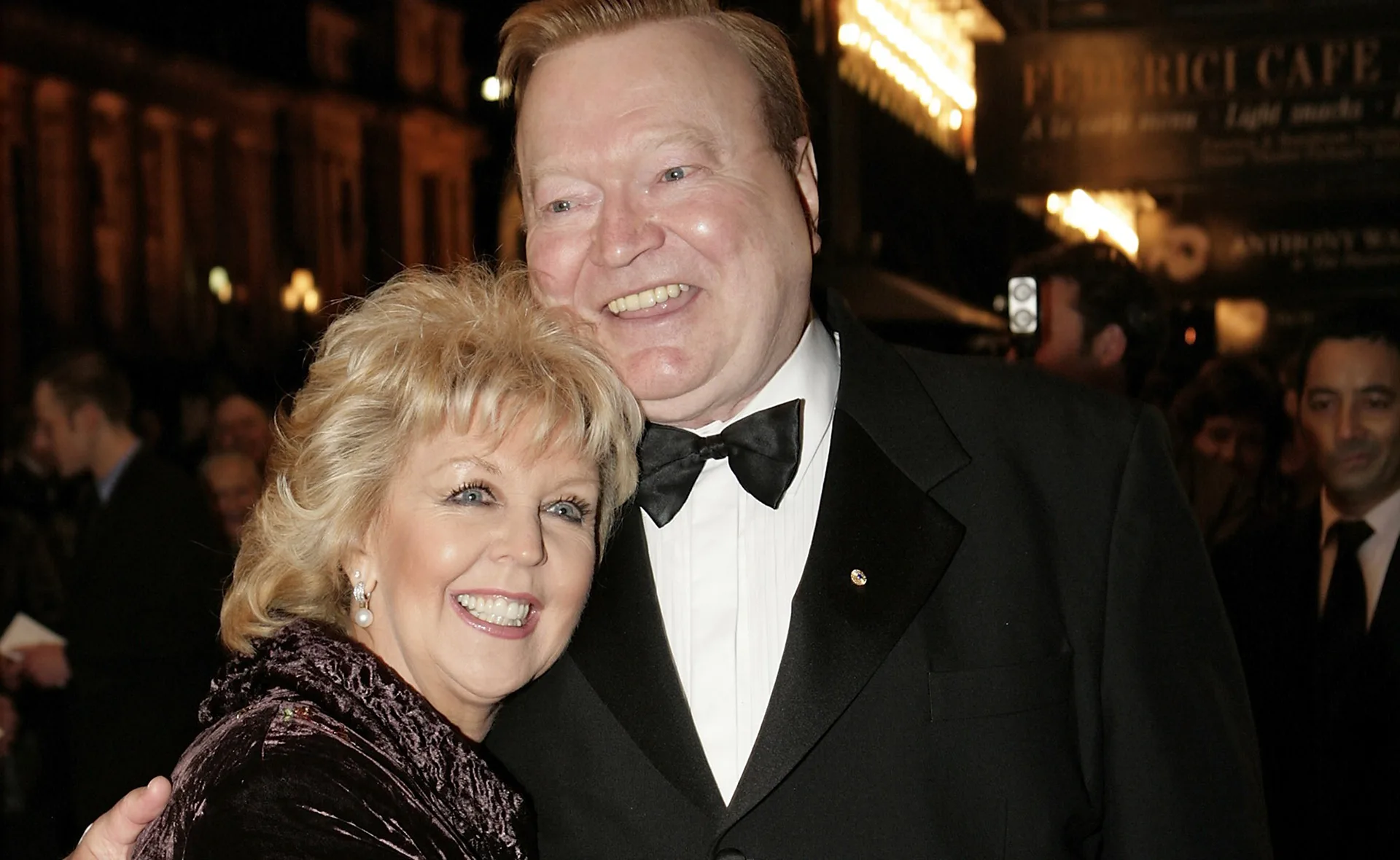 Couple in formal attire smiling at an event, with blurred crowd in background.