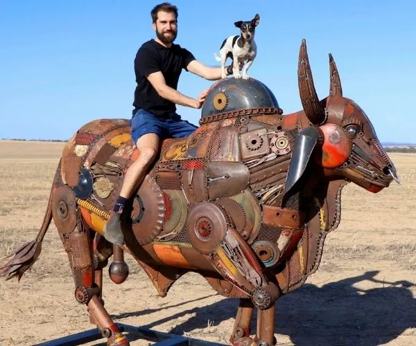 Man and dog on a robotic bull sculpture in a field under a clear blue sky.