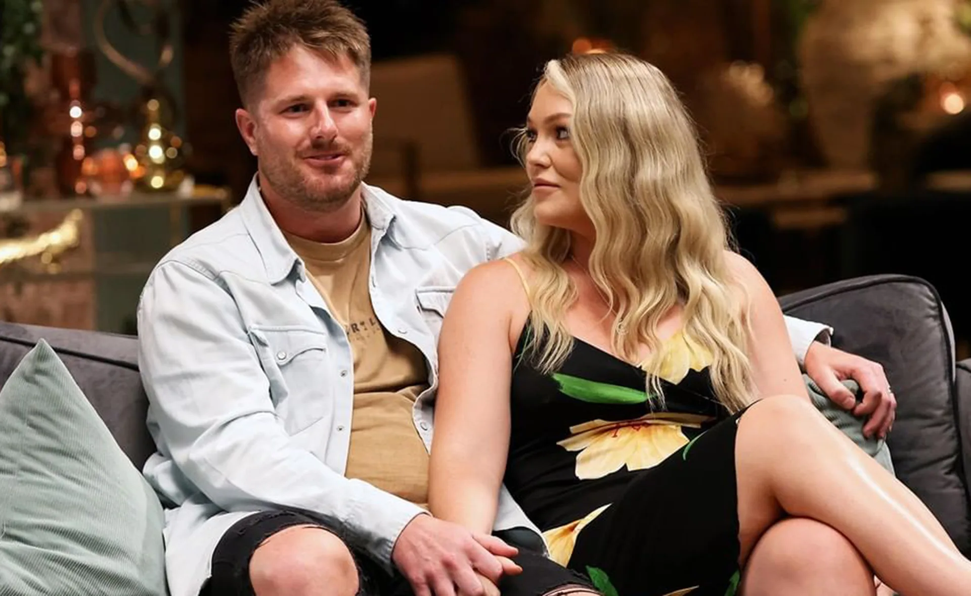A couple sitting on a couch, the man in a light shirt and the woman in a black floral dress, looking at each other.