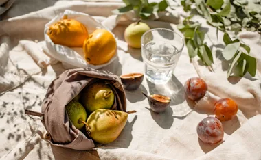 A picnic setup with pears, figs, lemons, plums, and a glass of water on a sunlit cloth.