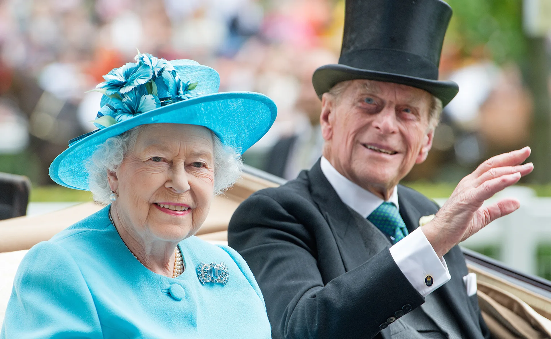 A woman in a light blue outfit and hat smiles, sitting beside a man in a black suit and top hat, waving at an event.
