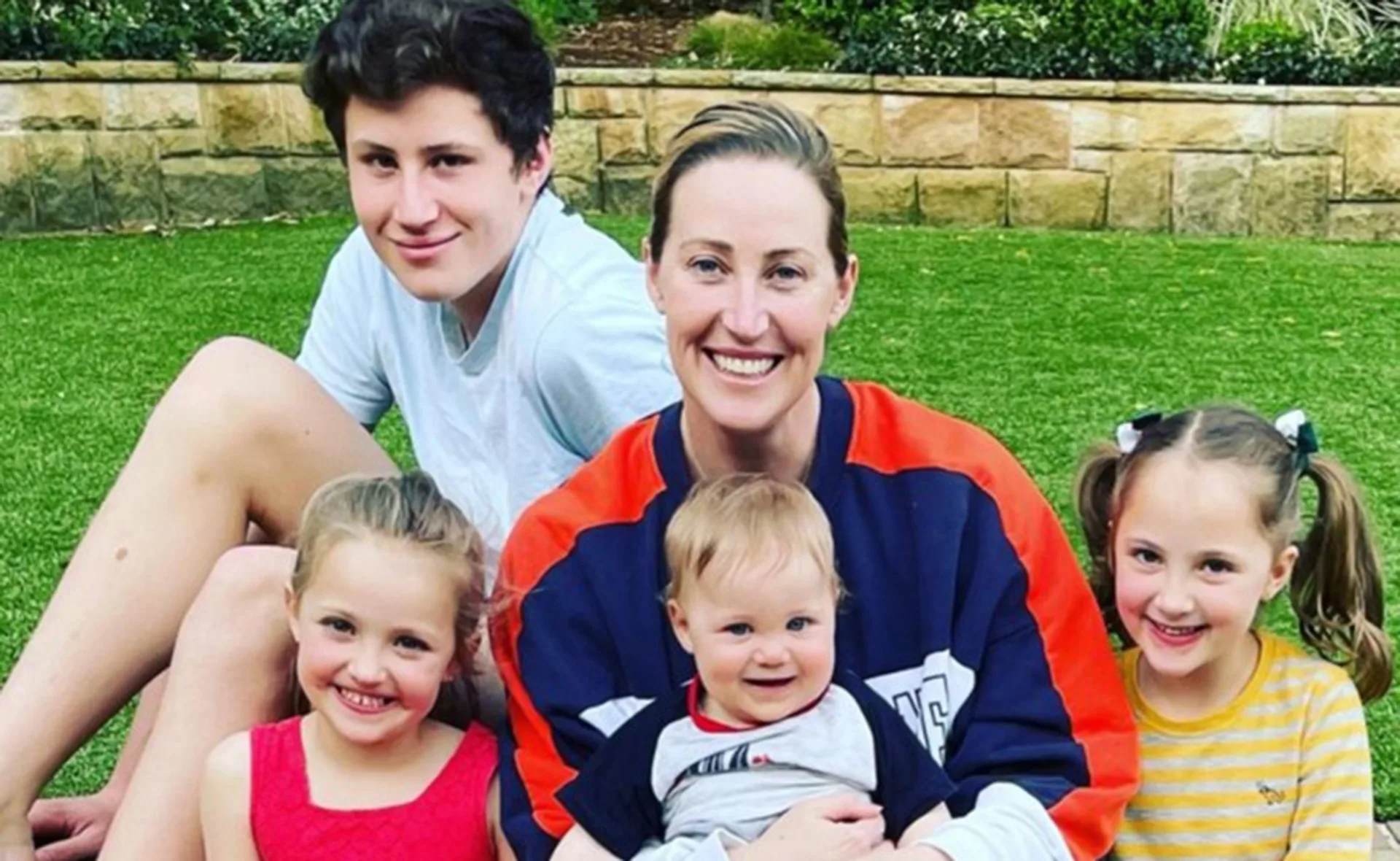 A smiling woman sits on grass with four children, two girls, one boy, and a baby, surrounded by greenery.