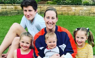A smiling woman sits on grass with four children, two girls, one boy, and a baby, surrounded by greenery.