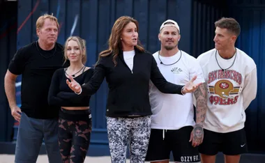 Five people standing outdoors, one in focus with arms outstretched. Casual attire, including a white shirt and sports sweatshirt.