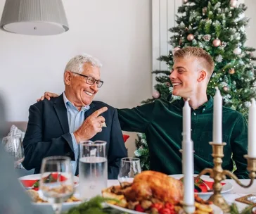 Grandfather and grandson laughing at a Christmas dinner table, with a decorated tree in the background.