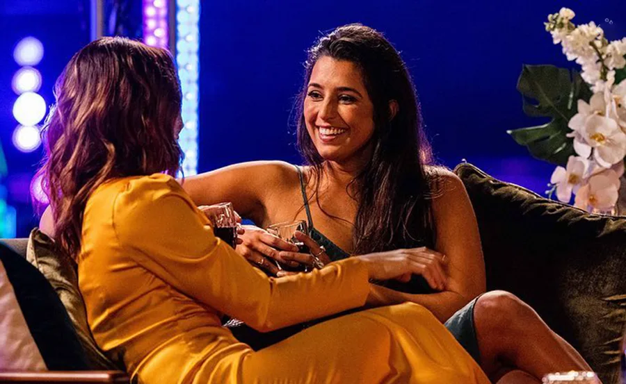 Two women sitting on a couch, one in gold dress, smiling and holding drinks against a colorful lit background.