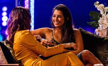 Two women sitting on a couch, one in gold dress, smiling and holding drinks against a colorful lit background.