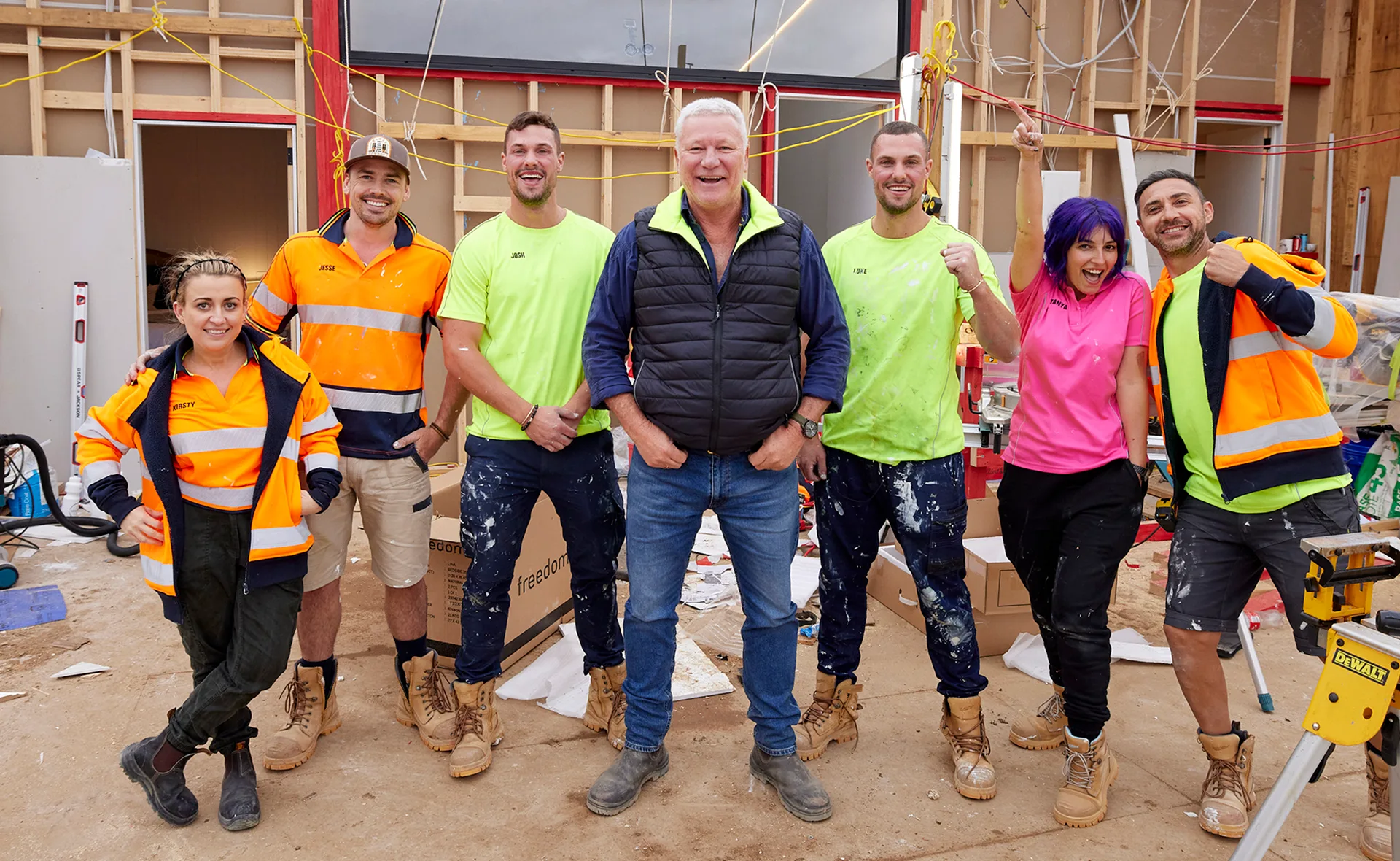 A group of seven construction workers smiling, wearing high-visibility clothing, on a building site with exposed walls and tools.