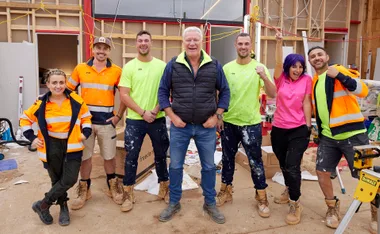 A group of seven construction workers smiling, wearing high-visibility clothing, on a building site with exposed walls and tools.