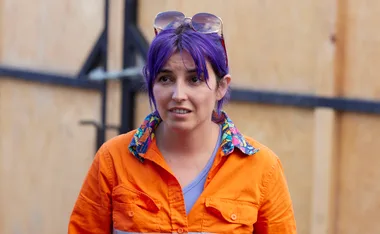 A person with purple hair and sunglasses on head wearing an orange shirt with a colorful collar, standing outdoors.