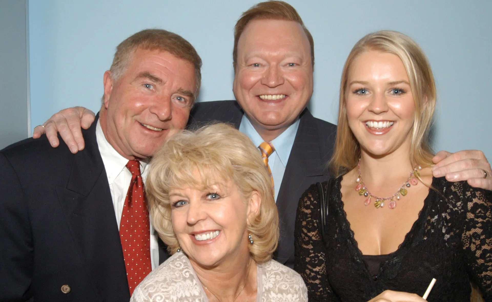 A group of four people smiling, including two men in suits, a woman in lace, and another woman in light clothing.