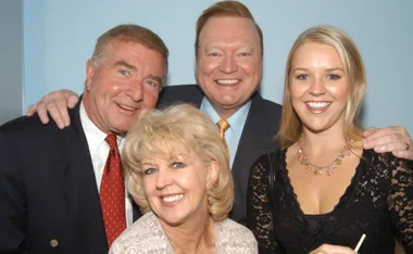 A group of four people smiling, including two men in suits, a woman in lace, and another woman in light clothing.