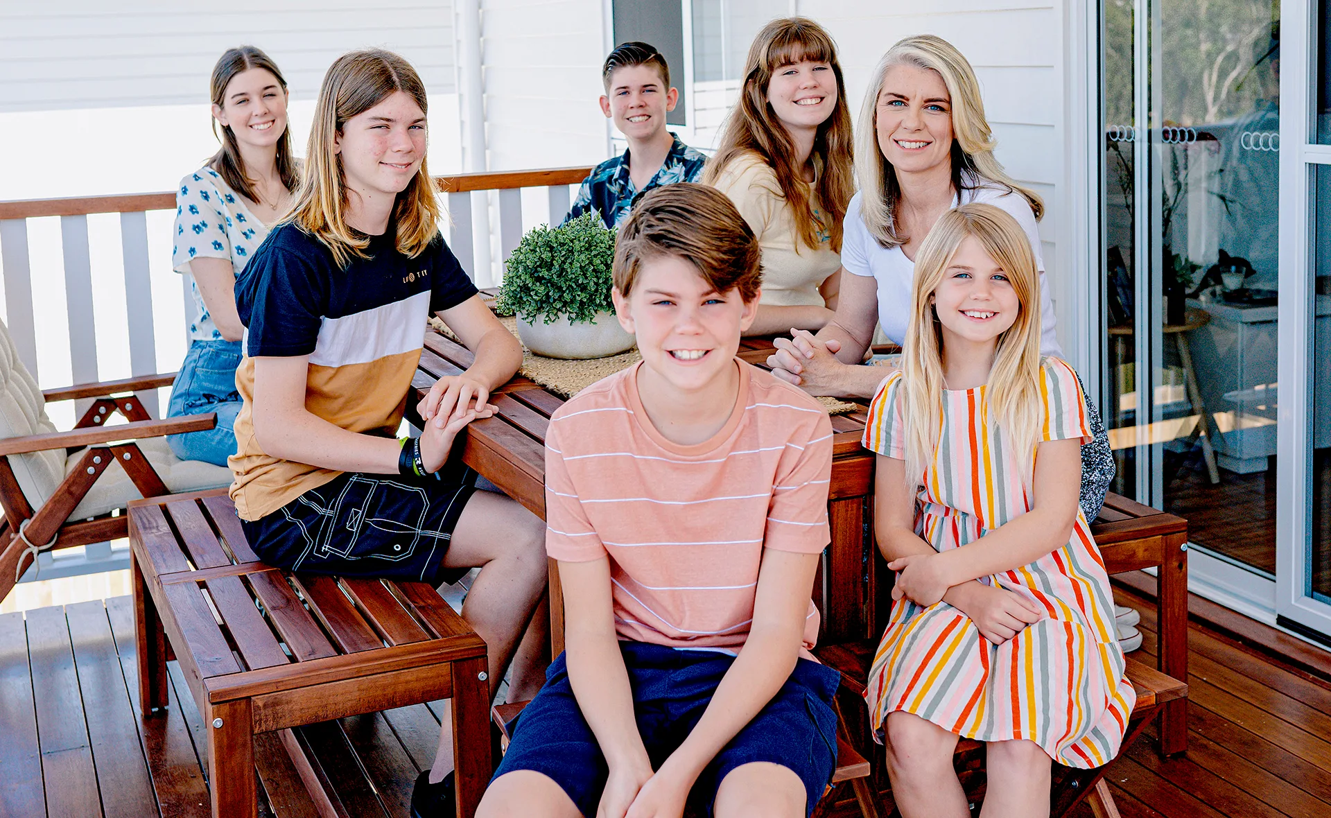 A family of seven smiling on a porch with wooden furniture and a plant centerpiece.