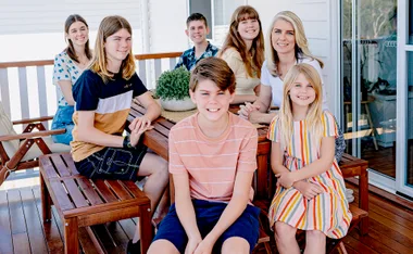 A family of seven smiling on a porch with wooden furniture and a plant centerpiece.
