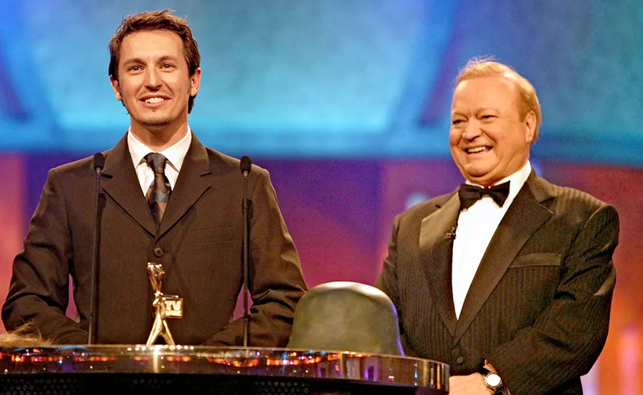 Two men in suits and ties stand at a podium with a TV award trophy, smiling during an award ceremony.