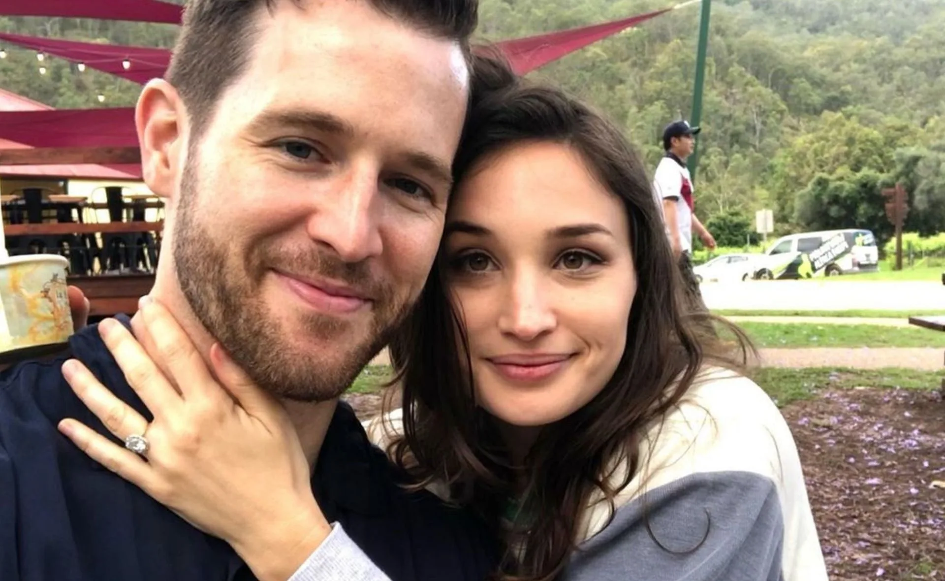 Couple smiling outdoors; woman shows engagement ring with hand on man's neck.