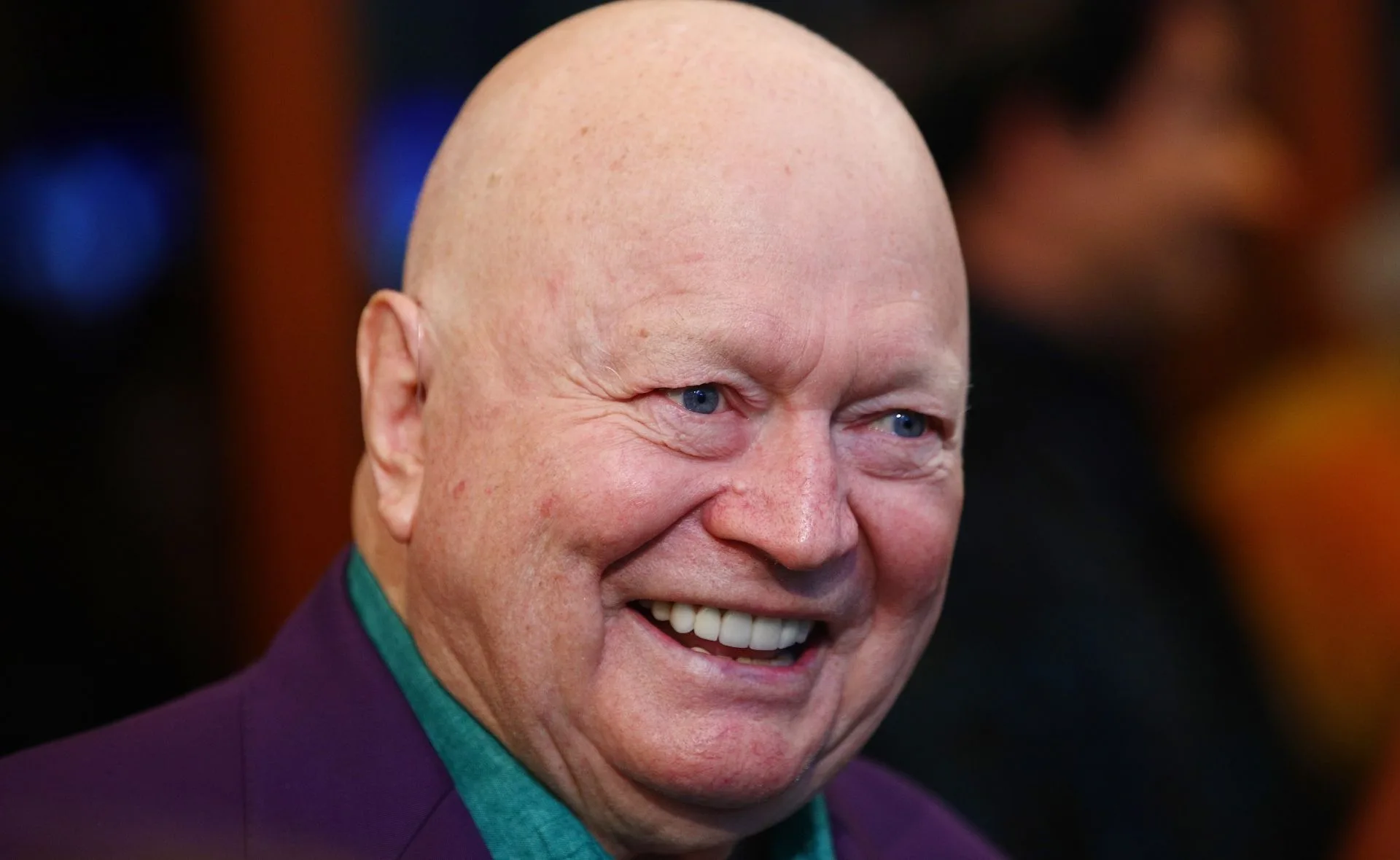 Smiling person with a bald head wearing a dark jacket and teal shirt in a close-up portrait.