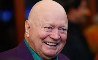 Smiling person with a bald head wearing a dark jacket and teal shirt in a close-up portrait.