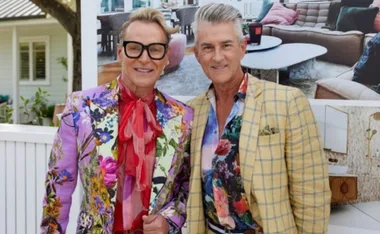 Two smiling men in vibrant, floral-patterned blazers, standing in front of a stylish living room interior.