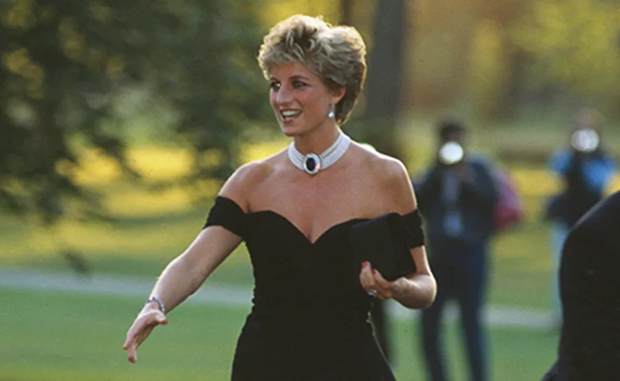 Princess Diana in a black off-shoulder dress with a white choker necklace, outdoors, smiling.