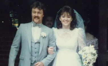 A couple stands arm in arm; the groom wears a grey suit and the bride a white dress, holding a bouquet.