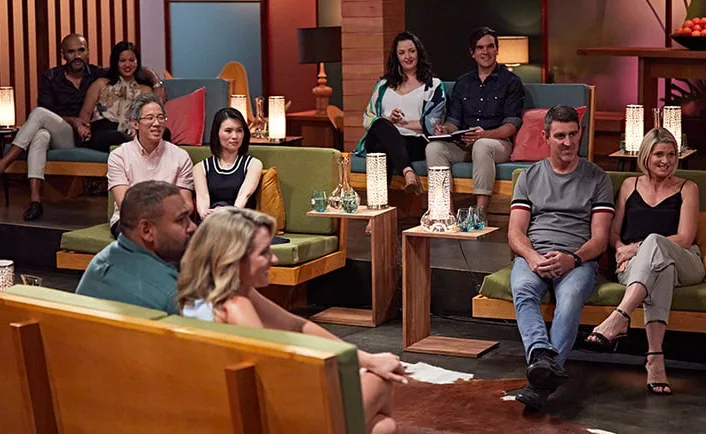 Group of people are seated in a modern room with warm lighting, attentively watching an event, some holding notebooks.