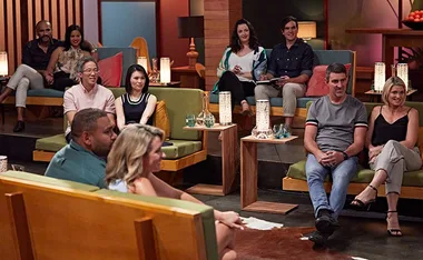 Group of people are seated in a modern room with warm lighting, attentively watching an event, some holding notebooks.