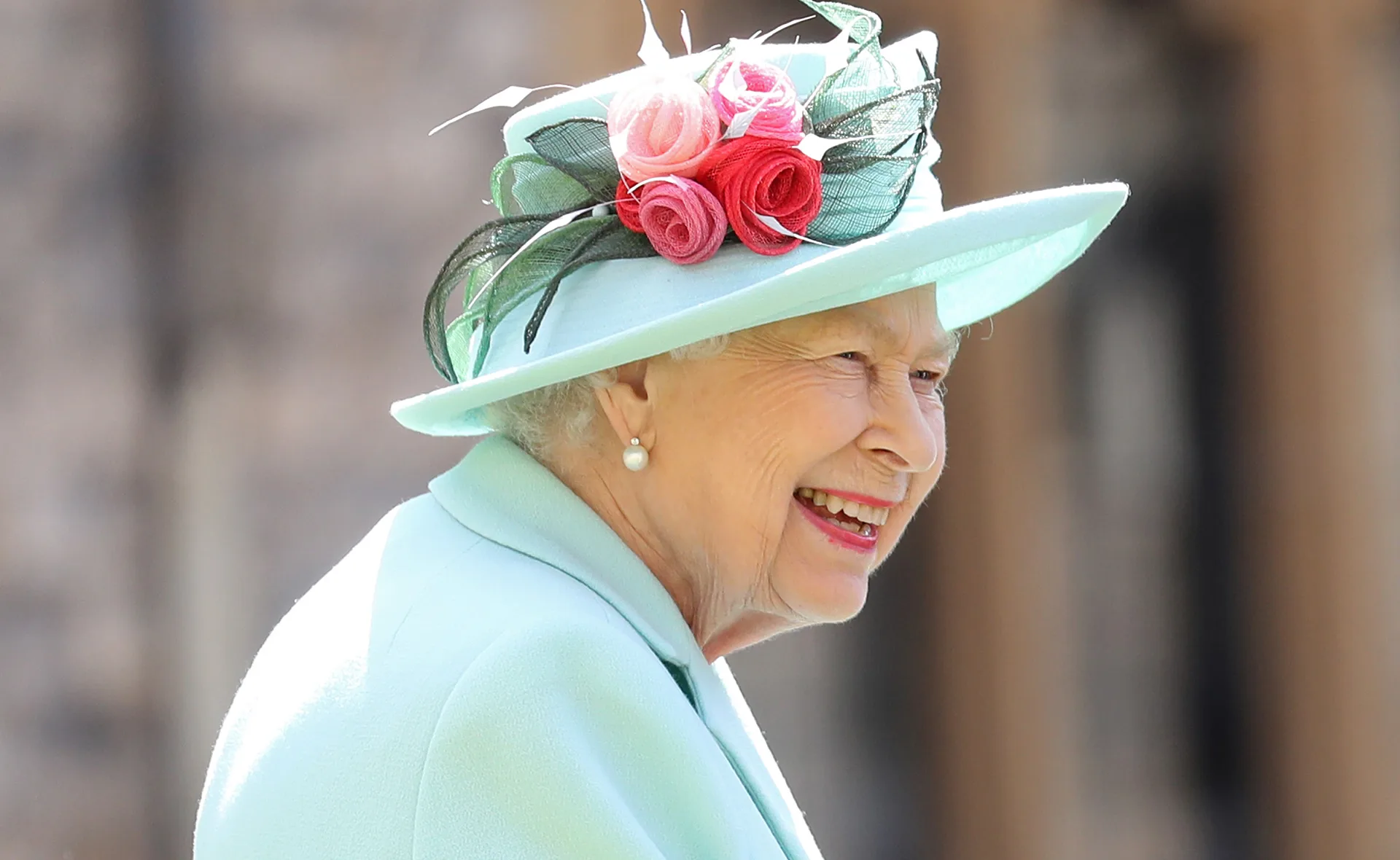 Elderly woman in a blue outfit, smiling, wearing a hat with pink flowers.