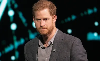 A man with red hair and a beard, wearing a gray blazer and shirt, in front of a blurred dark background.