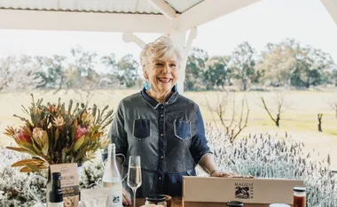 Elderly woman smiling by a table with flowers and gourmet products, in a rural setting.