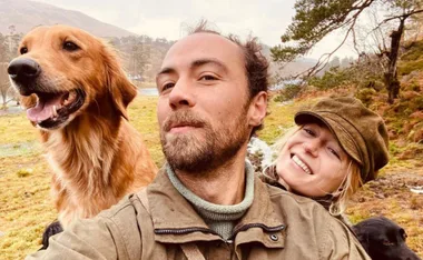 Man and woman in outdoor attire smiling with two dogs in a scenic, wooded landscape.