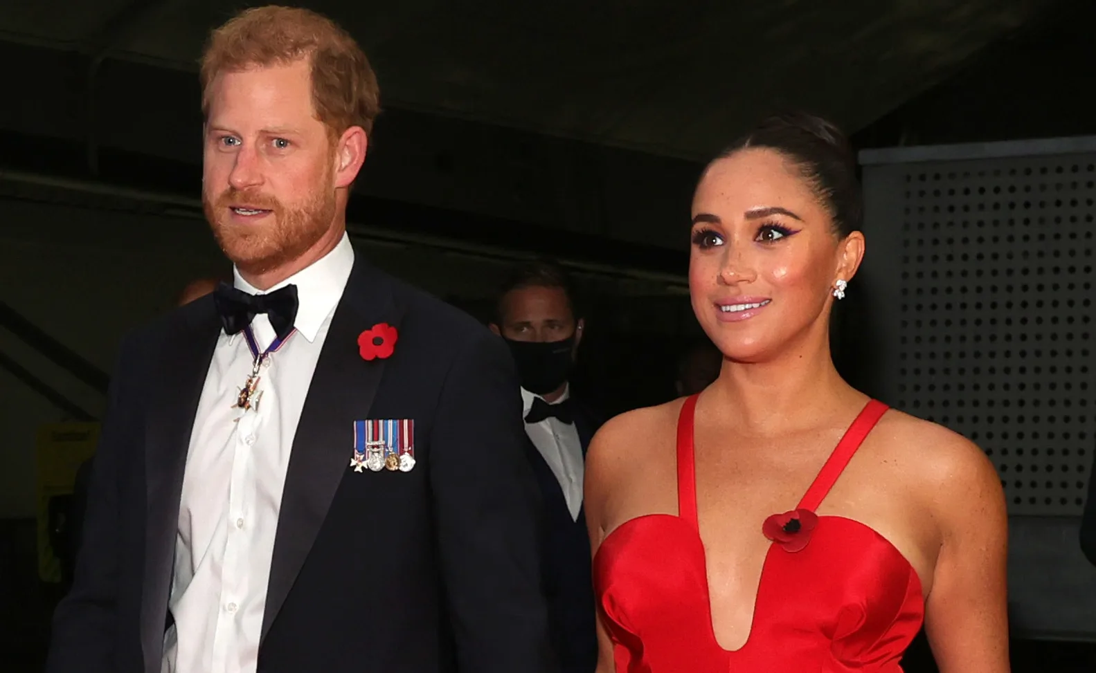 A man in a suit with medals and a woman in a red dress, both wearing poppies on their outfits at an event.
