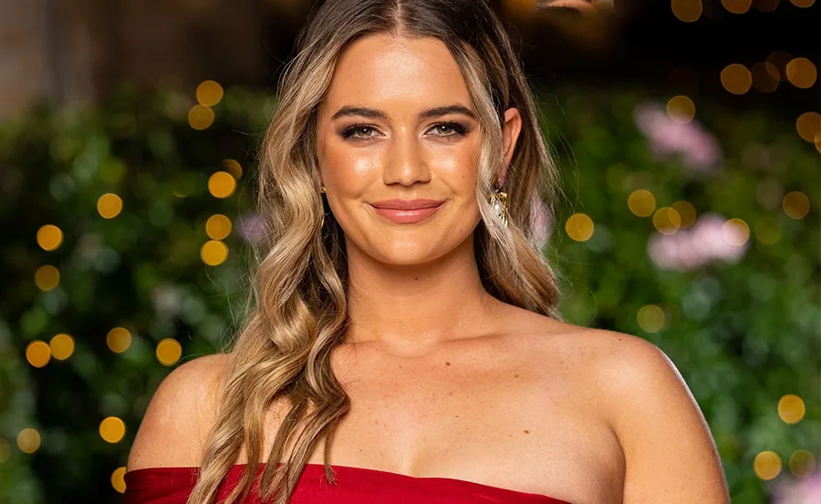A person in a red off-shoulder dress, smiling, with warm lighting and bokeh effect in the background.