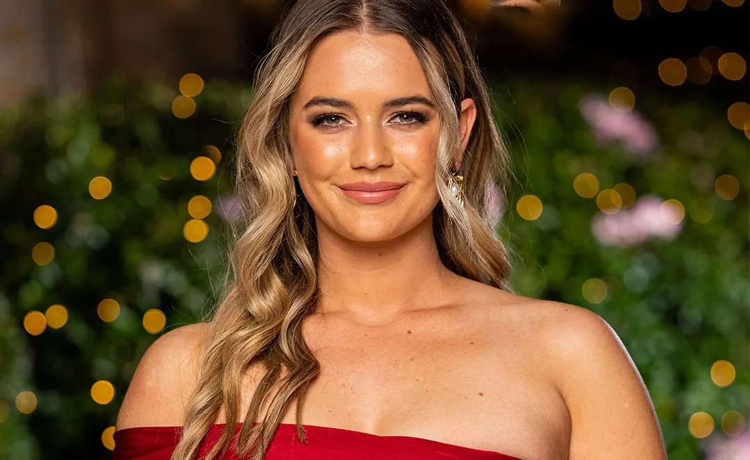 A person in a red off-shoulder dress, smiling, with warm lighting and bokeh effect in the background.