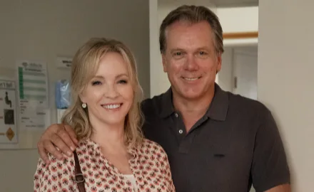 A man and woman smiling, standing closely indoors, with a hallway and wall signs in the background.