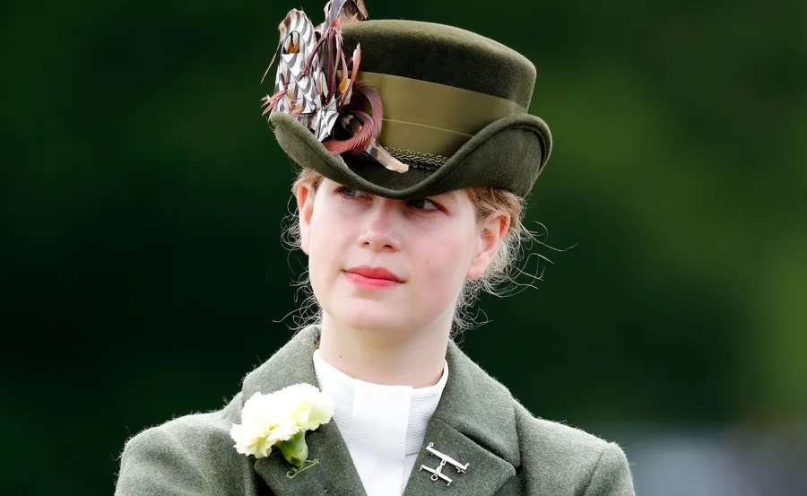 Young woman in a green hat adorned with feathers, wearing a green coat with a white flower pinned on it.